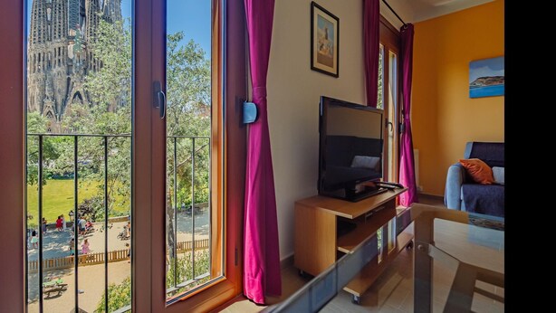 Habitación con televisor sobre un soporte de madera, un sofá y un balcón con vistas a un edificio en construcción.
