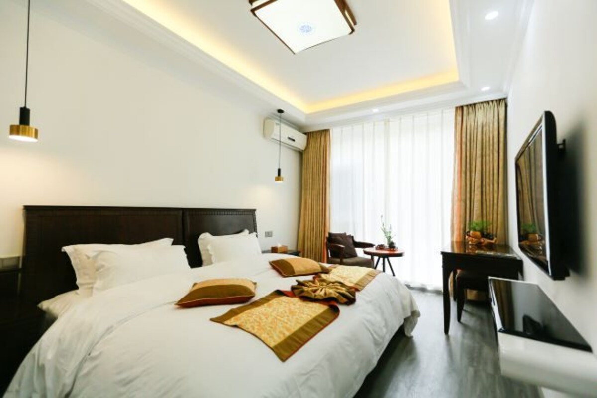 A modern hotel room with a large bed, bedside tables, a desk, a chair, a TV, and a ceiling light fixture.