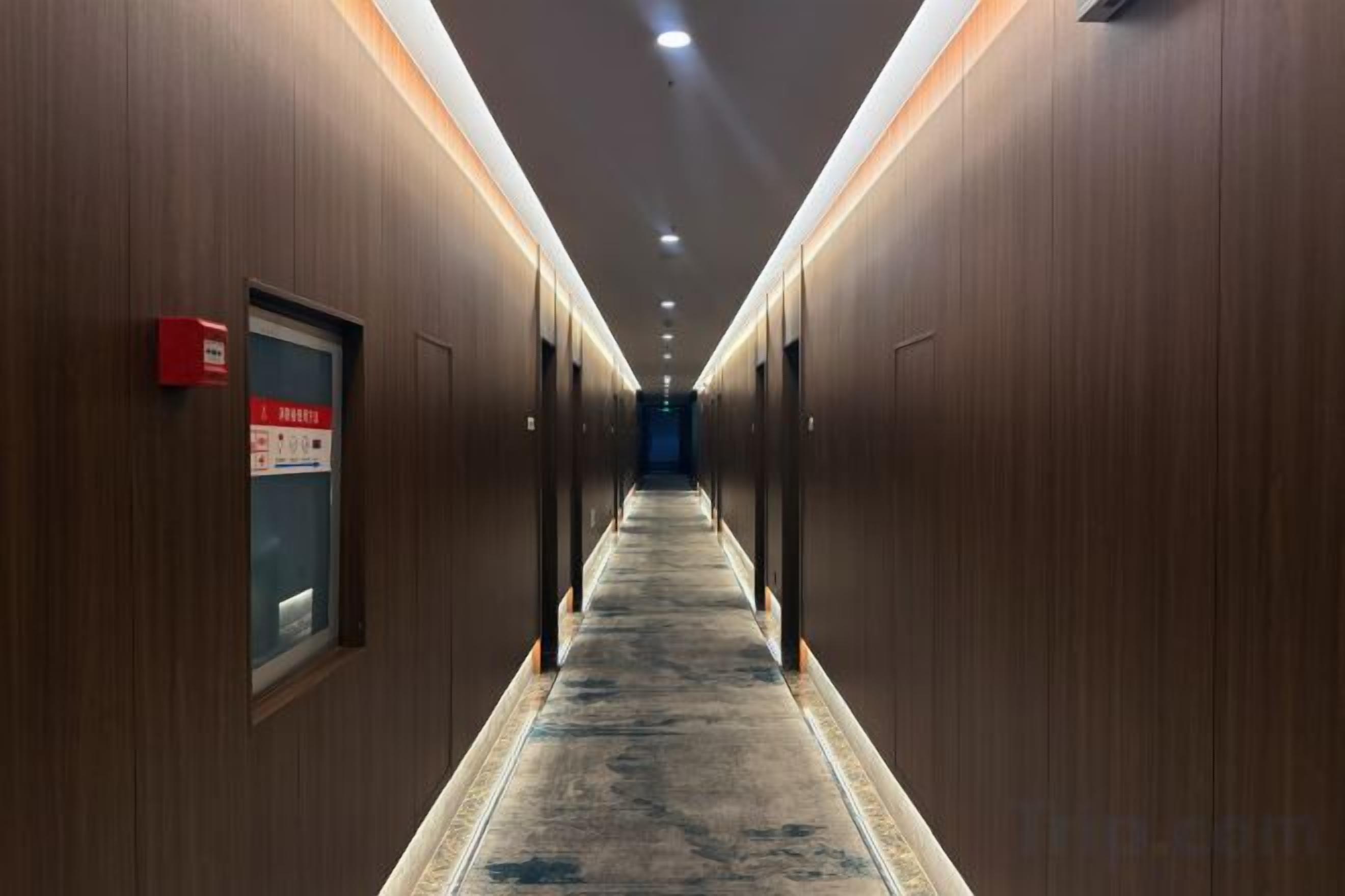 Corredor de hotel com paredes de madeira, piso coberto de carpete e placa de saída de emergência vermelha.