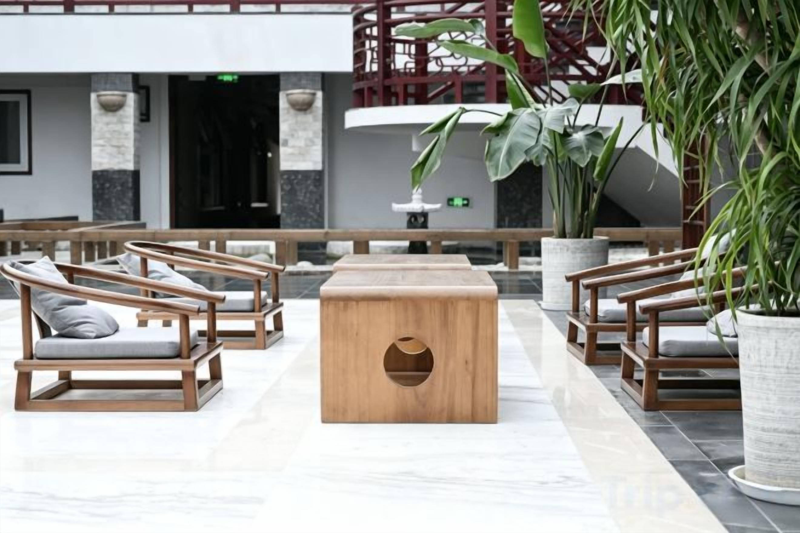 Um hall de entrada com cadeiras de madeira e uma mesa central, plantas em vasos, e uma escadaria ao fundo.