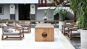 Um hall de entrada com cadeiras de madeira e uma mesa central, plantas em vasos, e uma escadaria ao fundo.