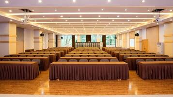 Un salón de conferencias con filas de mesas y sillas, un escenario al frente y un gran ventanal que proporciona luz natural.