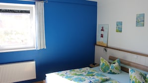 Quarto com parede azul, janela com cortinas brancas, cama com roupa de cama florida e uma pequena prateleira com objetos decorativos.