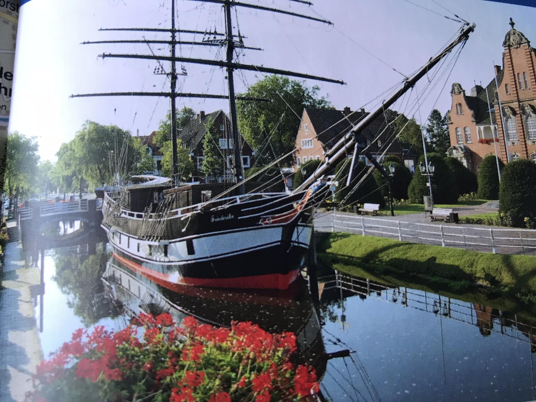 A traditional sailing ship docked at a canal, surrounded by historic buildings and lush greenery.
