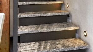 Un escalier avec des marches en granit et une rampe métallique, menant à un palier où se trouve un luminaire.