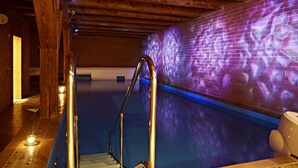 Un espace piscine avec un escalier menant à l’eau, un mur de briques sur lequel est projetée une lumière, et un plafond en bois.