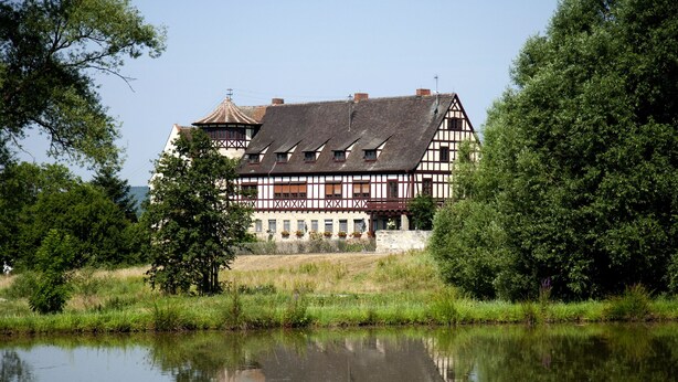 Un bâtiment traditionnel à colombages, avec un grand étang devant, entouré d’arbres.