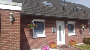 Una casa de ladrillo con puerta blanca, dos ventanas y un pequeño porche con macetas de flores.