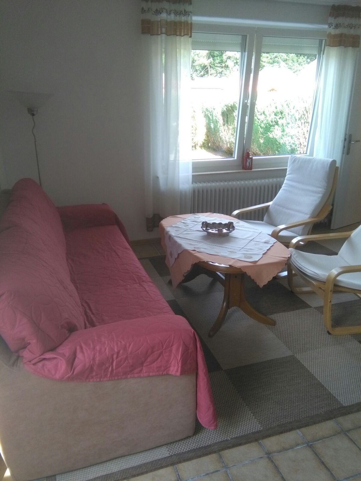 Ein Zimmer mit einer Couch, einem kleinen Tisch mit Unterlage, einem Sessel, einem Heizkörper und einem Fenster mit Vorhängen.