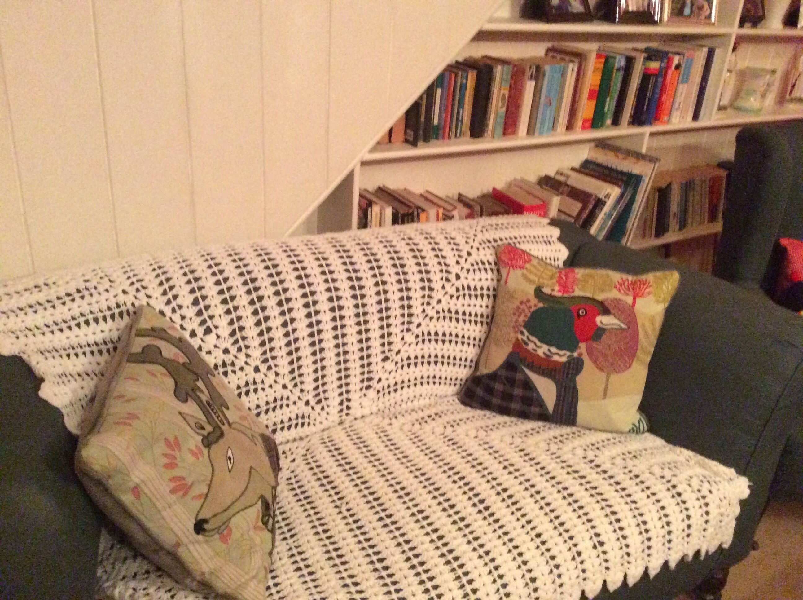 Ein gemütliches Wohnzimmer mit einem gehäkelten Sofa, einem Bücherregal voller Bücher und einem Zierkissen.