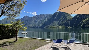 Una terraza con suelo de piedra, dos sillones azules, una sombrilla color beige y vistas a un lago con montañas al fondo.