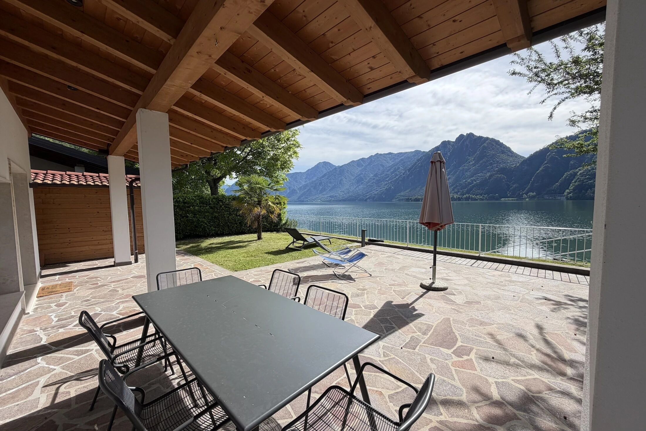 An outdoor patio with a table and chairs, a stone floor, a wooden ceiling, and a view of a lake and mountains.