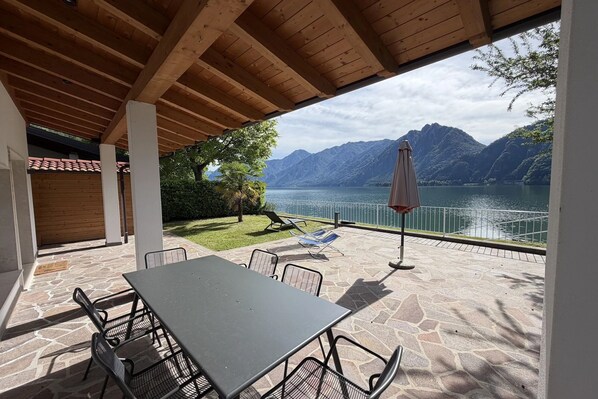 Un patio al aire libre con una mesa y sillas, suelo de piedra, techo de madera y vistas a un lago y montañas.