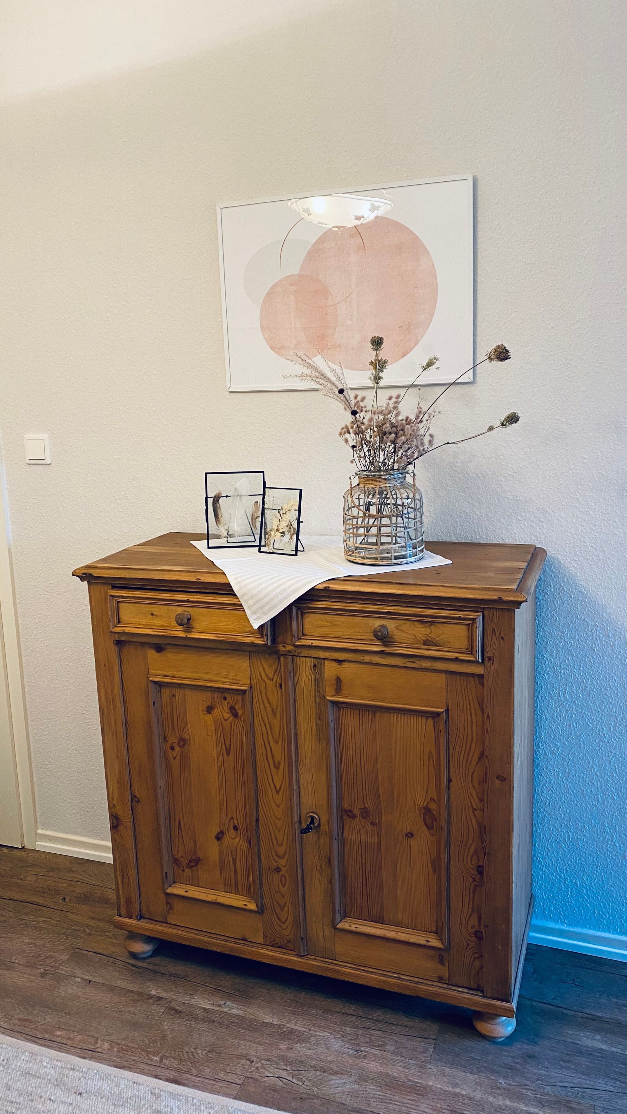 Ein Holzkabinent mit zwei Schubladen, eine Vase mit getrockneten Blumen und ein gerahmtes Kunstwerk an der Wand.