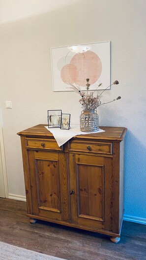 Un mueble de madera con dos cajones, un jarrón con flores secas y una obra de arte enmarcada en la pared.