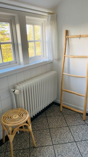 Ein Zimmer mit Heizkörper, einem Korbstuhl, einer Holzleiter und einem Fenster mit weißen Vorhängen.