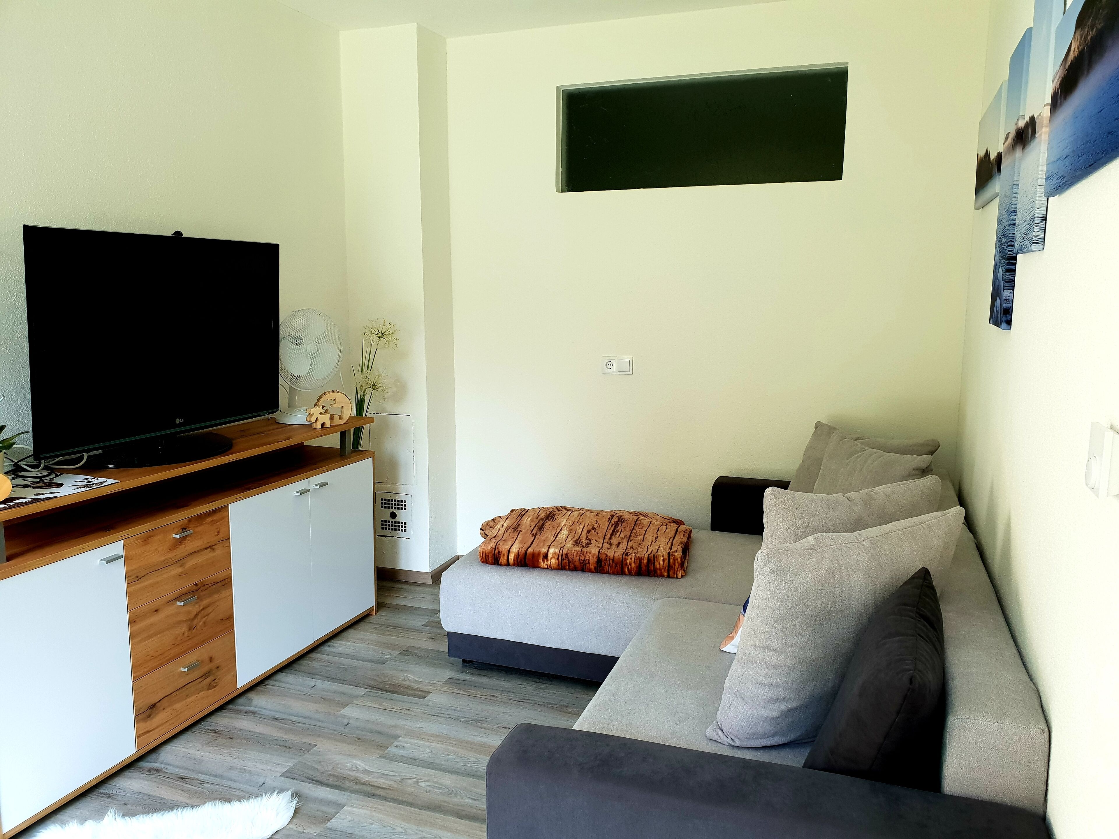 A modern living room with a flat-screen TV, wooden cabinet, grey sofa, and a small table with decorative items.