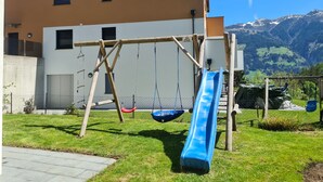 A playground with a slide and swings in front of a building with a mountain view. - Apartment in a quiet location (Fügenberg)