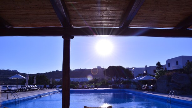 Zona de piscina con sillones de descanso y sombrillas, cielo azul despejado y vista de edificios al fondo.