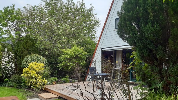 Una casa con techo inclinado, una terraza de madera y un jardín con diversas plantas y árboles.