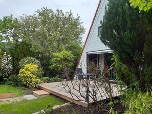 Una casa con techo inclinado, una terraza de madera y un jardín con diversas plantas y árboles.