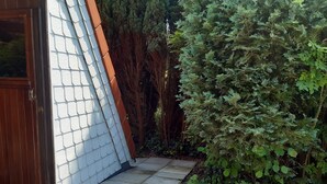 Una terraza de madera con un edificio de revestimiento blanco, una puerta de madera y un seto verde frondoso.