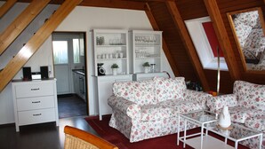 Una habitación de ático acogedora con techo de madera, un conjunto de sofá blanco, una mesa de centro de vidrio y una silla de mimbre. Hay armarios blancos con puertas de vidrio y una ventana con marco rojo.