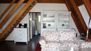 Un dormitorio en el ático con techo de madera, un sofá floral, un aparador blanco con cristalería y una alfombra roja.