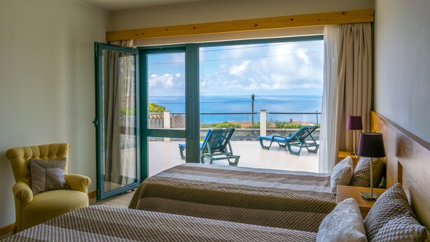 Habitación de hotel con cama, una silla, una mesita de noche y vista al océano a través de un ventanal grande.