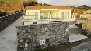 Una casa de dos pisos con un muro de piedra en el frente, techo de tejas y un balcón con barandillas de cristal.