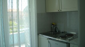 Una pequeña cocina con un refrigerador blanco, una estufa y un lavamanos. Hay una ventana con cortinas translúcidas y vistas a edificios.