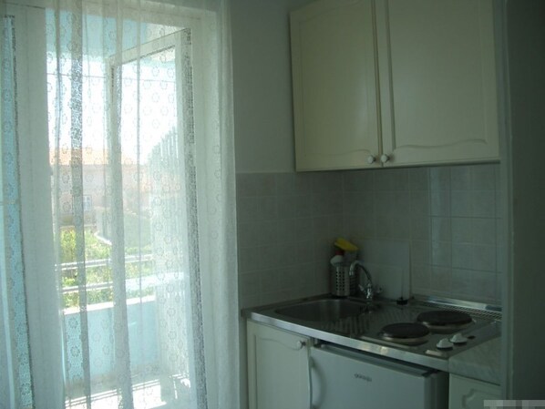 A small kitchen with a white refrigerator, a stove, and a sink. There is a window with sheer curtains and a view of buildings outside.