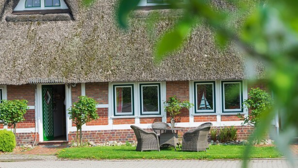 A thatched-roof cottage with a brick facade, a green door, and a small garden with wicker furniture.