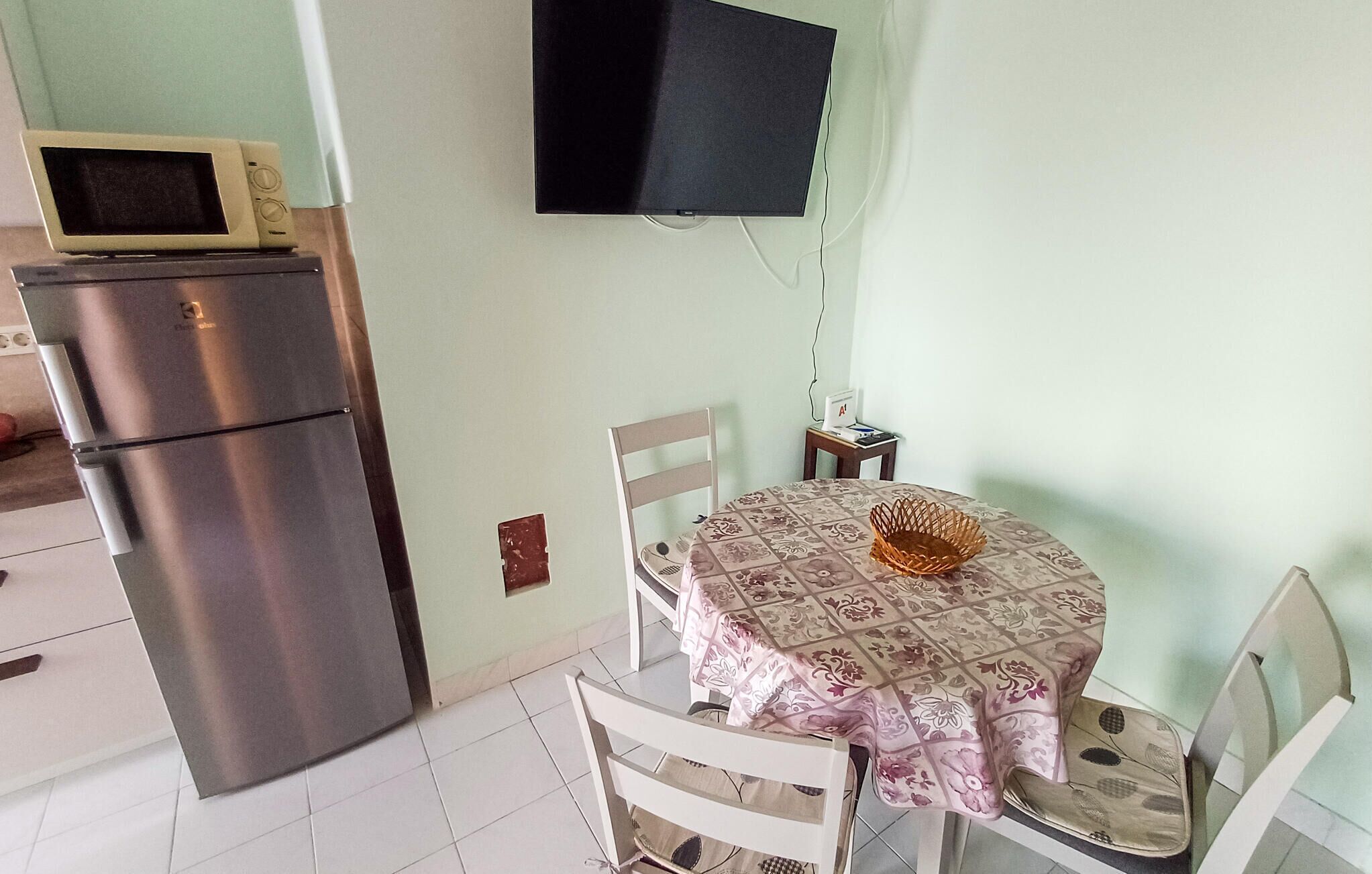 A compact kitchen with a refrigerator, a microwave, a dining table with chairs, and a flat-screen TV mounted on the wall.