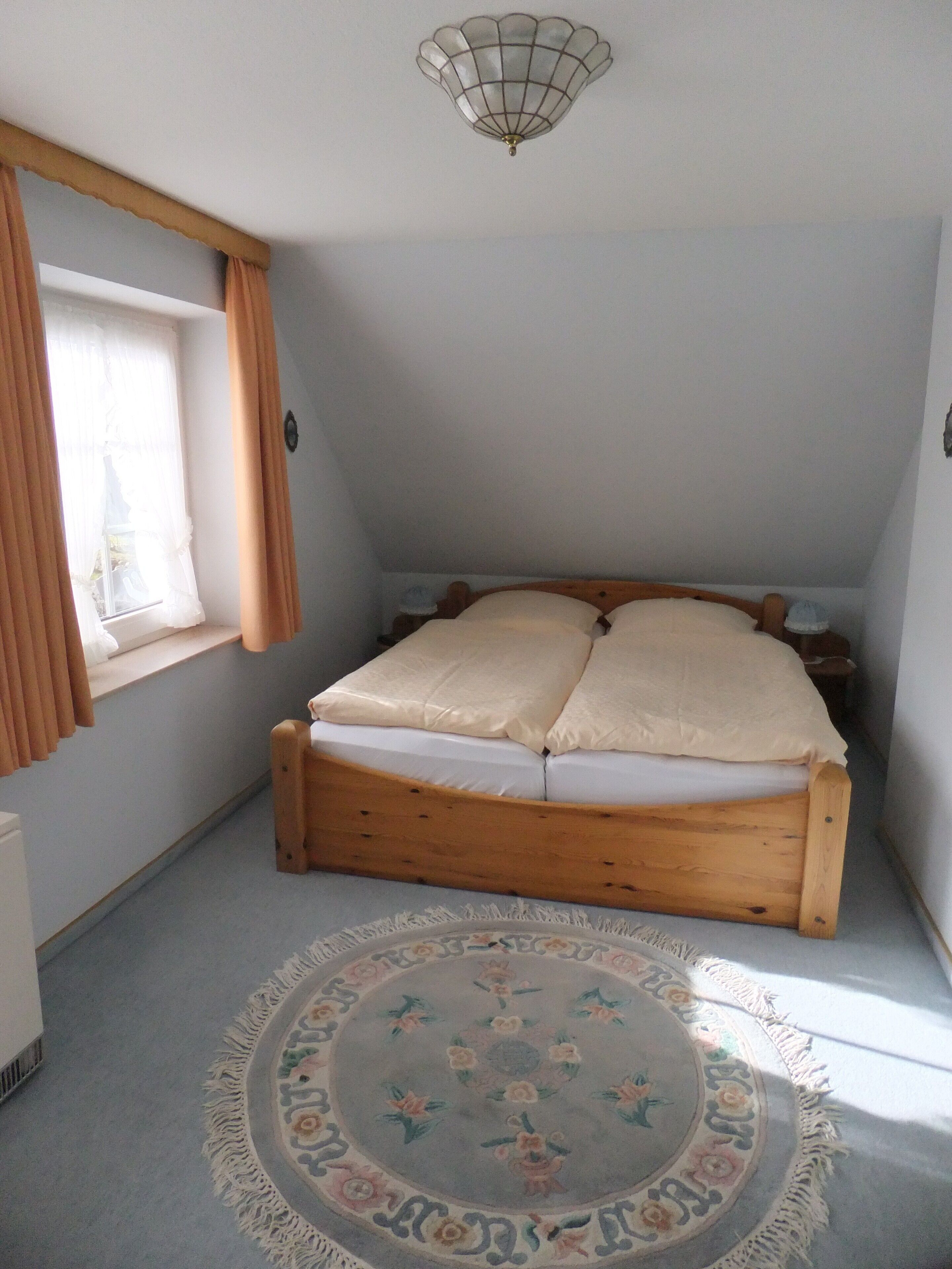 Une chambre à coucher avec un lit en bois, un tapis rond à motifs floraux, une fenêtre avec des rideaux et un luminaire au plafond.