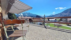 Un balcón con banca de madera, baranda metálica y vista a un paisaje montañoso con casas y un telesilla.