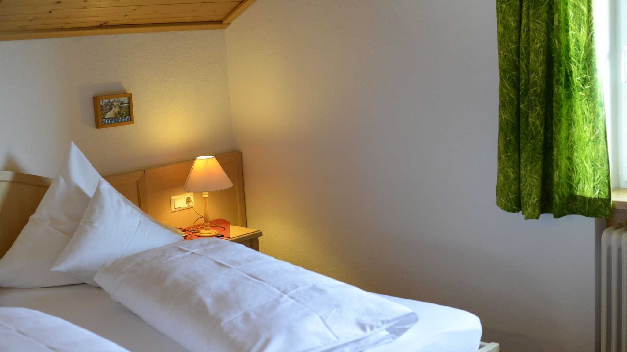 A neatly made bed with white linens, a bedside lamp, a framed picture on the wall, and a window with green curtains.