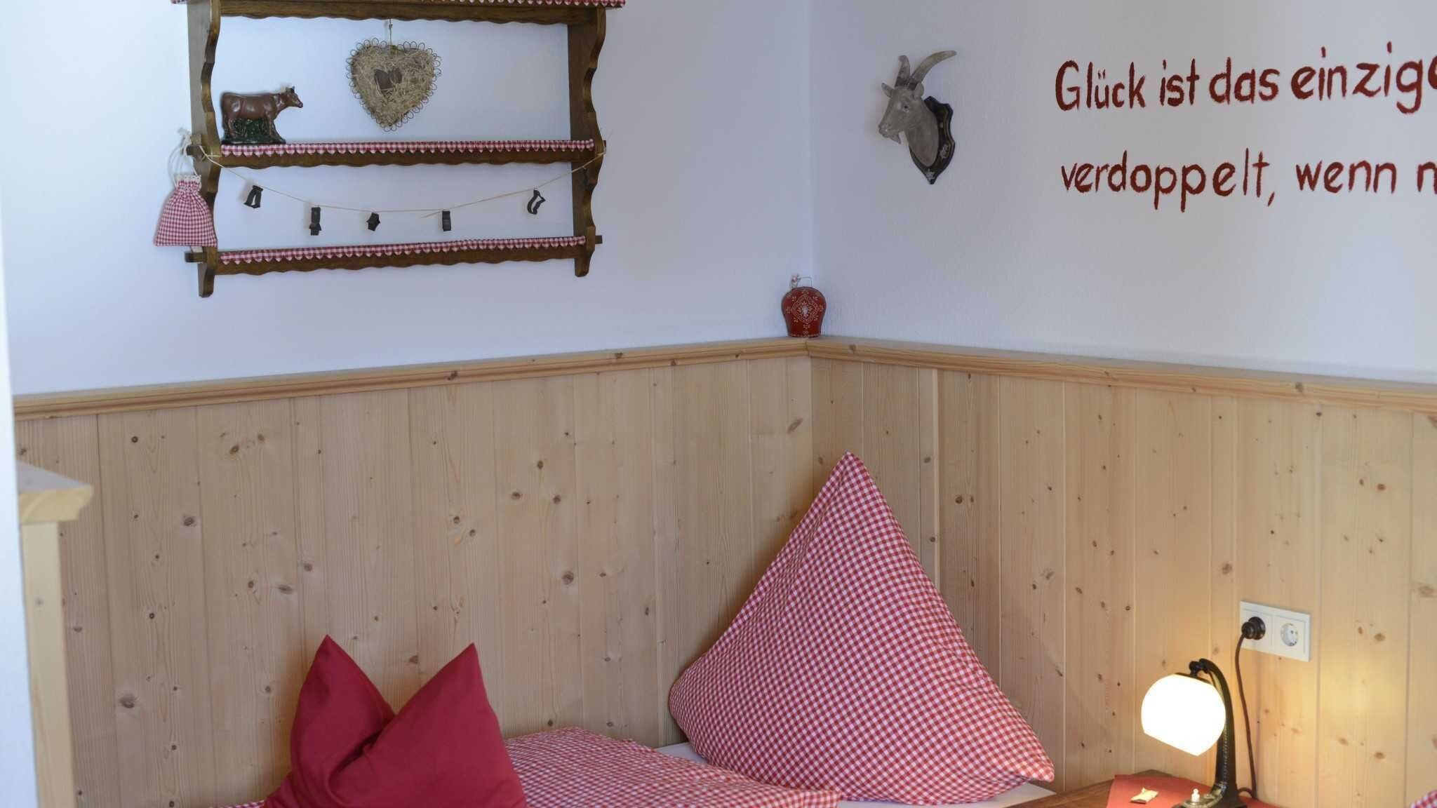 Ein Schlafzimmer im Eckbereich mit Holzvertäfelung, einem Bett mit rot-weiß karierter Bettwäsche, einem an der Wand befestigten Regal mit Dekoration und einer Wandleuchte.