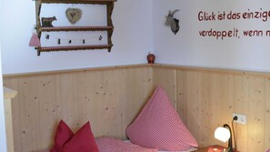 Une chambre d’angle avec un lambris en bois, un lit recouvert d’une couette à carreaux rouges et blancs, une étagère murale ornée d’objets décoratifs et une lampe fixée au mur.