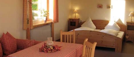 Un dormitorio con cama, mesitas de noche, una mesa con mantel a cuadros y un ventanal con cortinas.