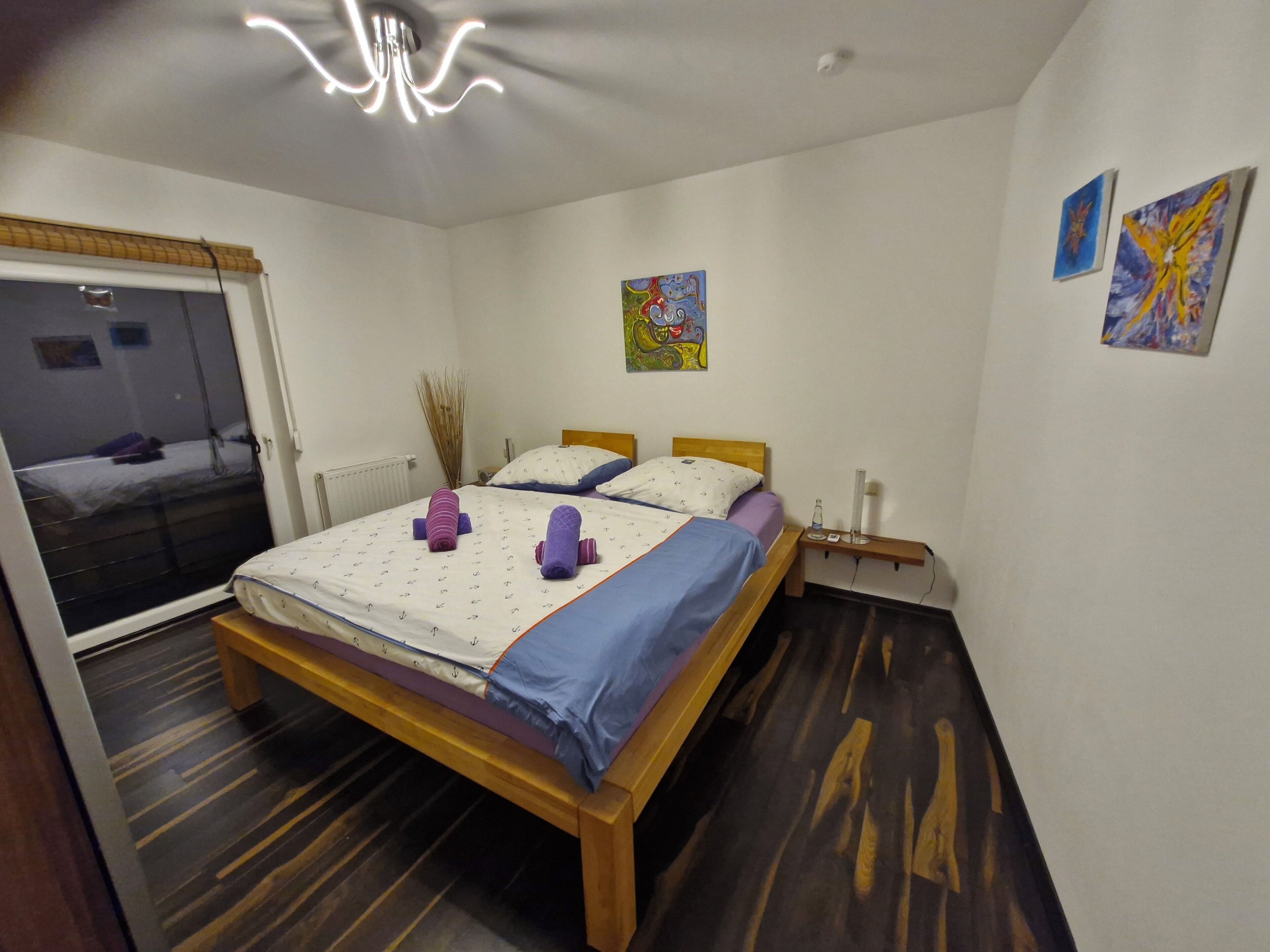 A bedroom with a wooden bed, a window with blinds, a bedside table with a lamp, and artwork on the wall.