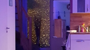 Een kamer met een houten trap, een deur met een gouden glinsterende gordijn, een witte kast en een kleine tafel met een fles en een beker.