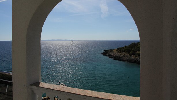 A view through an arched window of a coastal area with a sailboat on the water and rocky terrain on the right. - Beach Villa: private bathing area, sandy beach, WiFi, ideal for families (Novalja)