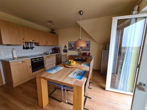 A compact kitchen with wooden cabinets, a dining table with placemats, a hanging light fixture, and a view through a glass door.