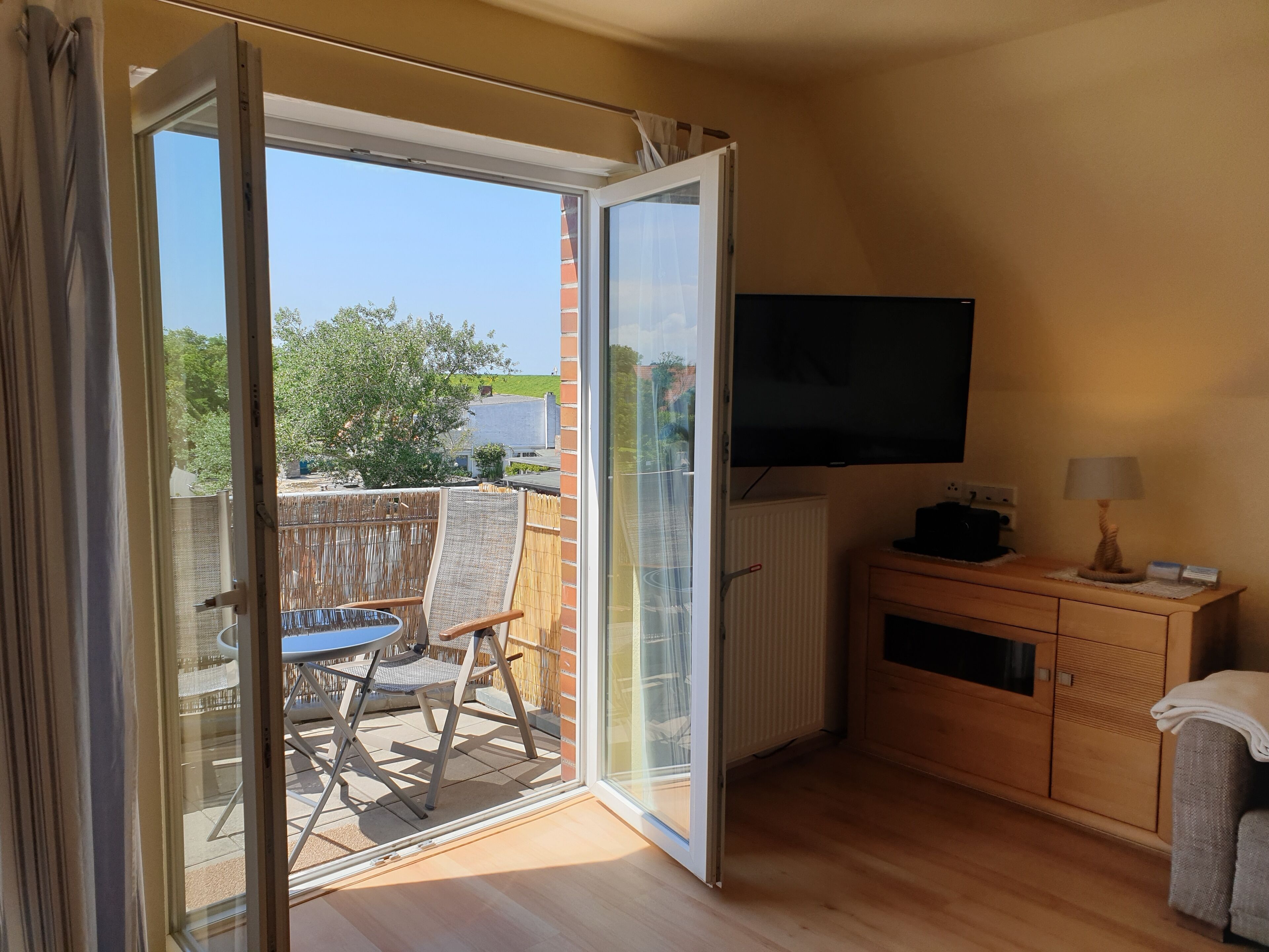 A room with a sliding glass door leading to an outdoor area with a table and chairs. A flat-screen TV is mounted on the wall, and there is a wooden cabinet with a lamp on top.