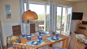 Een eetgedeelte met een tafel gedekt voor vier personen, een rieten stoel, een blauwe met kussens beklede bank en een televisie op de achtergrond.
