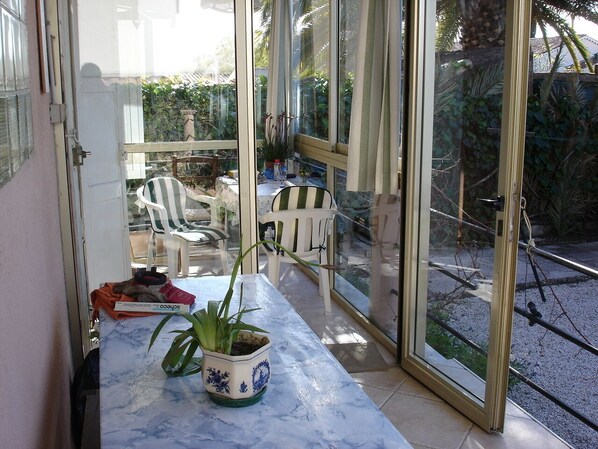 A glass-enclosed patio with a white table, two potted plants, and a white chair.