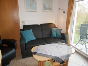 A living room with a dark grey sofa, two teal cushions, a wooden coffee table with a patterned tablecloth, a black leather chair, and a radiator.