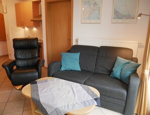 A living room with a leather chair, a grey sofa with blue cushions, and a small wooden table with a patterned tablecloth.