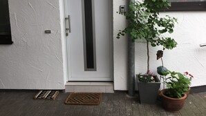 Une porte blanche avec une bande noire verticale, un pot de fleurs contenant des fleurs rouges et un petit nichoir fixé à un mur blanc.